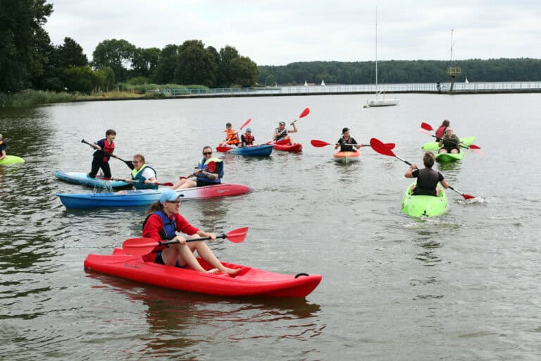 Dziewięć różnokolorowych kajaków na wodzie. Płyną w nich w różne strony młodzi ludzie. W tle pomost, za nim, na horyzoncie linia lasu