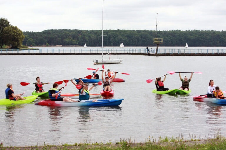 Na pierwszym planie pas trawy, tuż za nim zbiornik wodny i kilka kolorowych kajaków. Niektóre osoby z kajaków unoszą nad głowę wiosła. W tle pomost, za nim na linii horyzontu las
