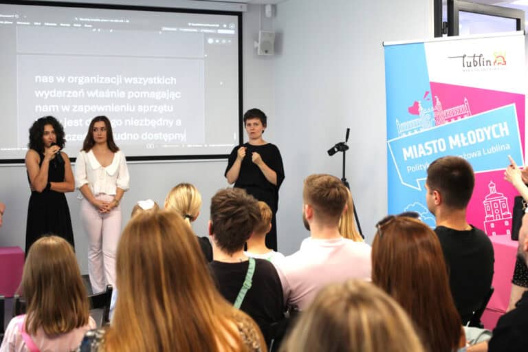 Na pierwszym planie ludzie siedzący tyłem do obiektywu. Plan drugi od lewej: kobieta w czarnej sukience, trzymająca mikrofon przy ustach, obok autorka prezentowanych fotografii. Od prawej tłumaczka języka migowego, obok baner stojący z napisem: Miasto Młodych. W tle monitor wyświetlający napisy dla osób słabosłyszących