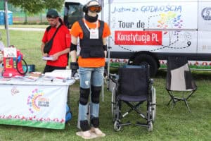 Stoisko Tour de Konstytucja, Jestem za Godnością. Od lewej mężczyzna trzymający w rękach broszury. W centrum kadru symulator starości (manekin wielkości człowieka, w goglach i słuchawkach i kamizelce). Tuż przy nim stoi wózek dla osób z niepełnosprawnością