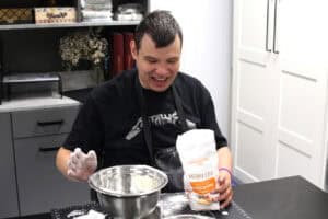 W centralnej części kadru śmiejący się mężczyzna. Przed nim metalowa miska i torba z mąką. Mężczyzna trzyma ją w lewej dłoni, prawą dłoń trzyma nad miską, jest cała w mące. Za nim regał biurowy, z prawej fragment szafy kuchennej