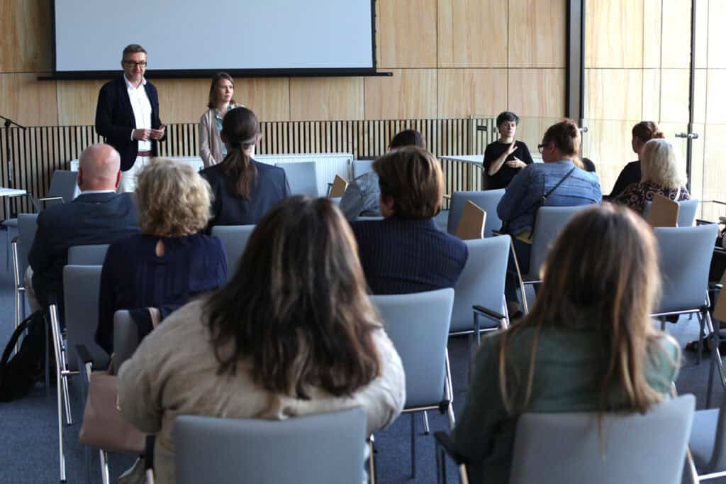 Przed rzędami krzeseł z siedzącymi na nich ludźmi stoją mężczyzna w okularach, obok niego kobieta. Z prawej siedzi tłumaczka języka migowego. Za nimi dolna część ekranu wiszącego na ścianie z drewnianych paneli; widok z perspektywy widowni