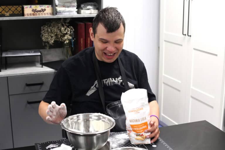 W centralnej części kadru śmiejący się mężczyzna. Przed nim metalowa miska i torba z mąką. Mężczyzna trzyma ją w lewej dłoni, prawą dłoń trzyma nad miską, jest cała w mące. Za nim regał biurowy, z prawej fragment szafy kuchennej