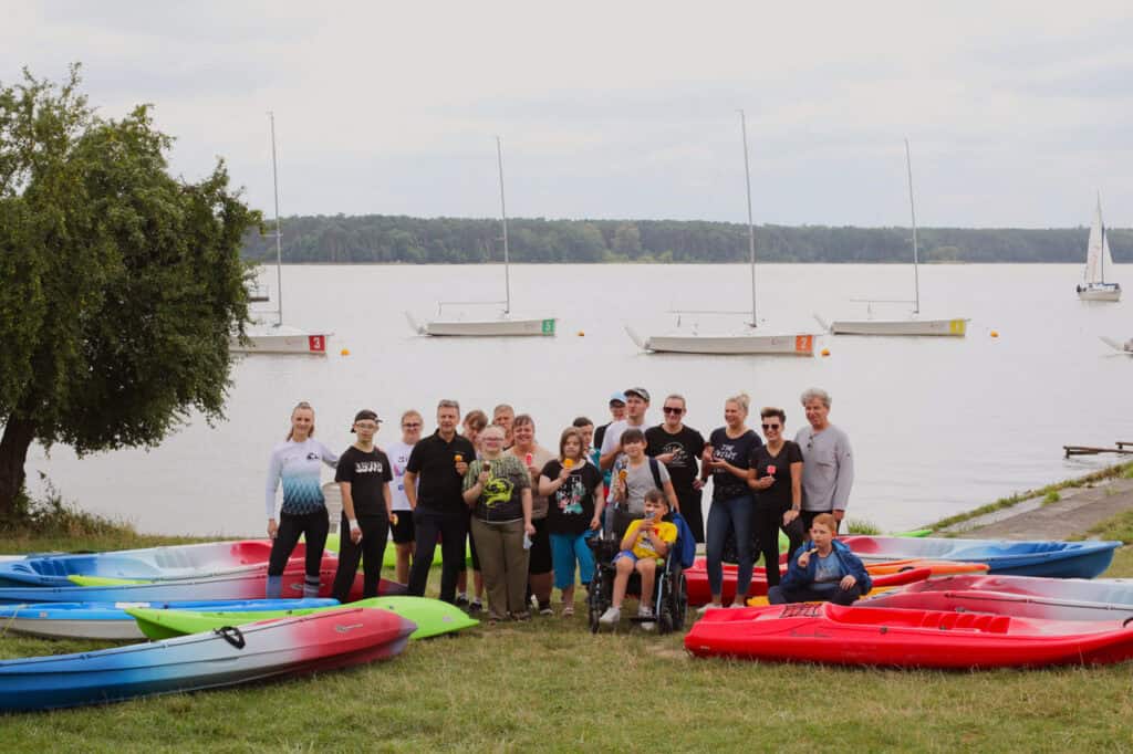 Na pierwszym planie między rozłożonymi na trawie kolorowymi kajakami stoi grupa kilkunastu osób pozujących do zdjęcia. Niektórzy trzymają w dłoniach lody na patyku. Przed nimi chłopiec na wózku. W tle zbiornik wodny i kilka żaglówek. Na horyzoncie linia lasu