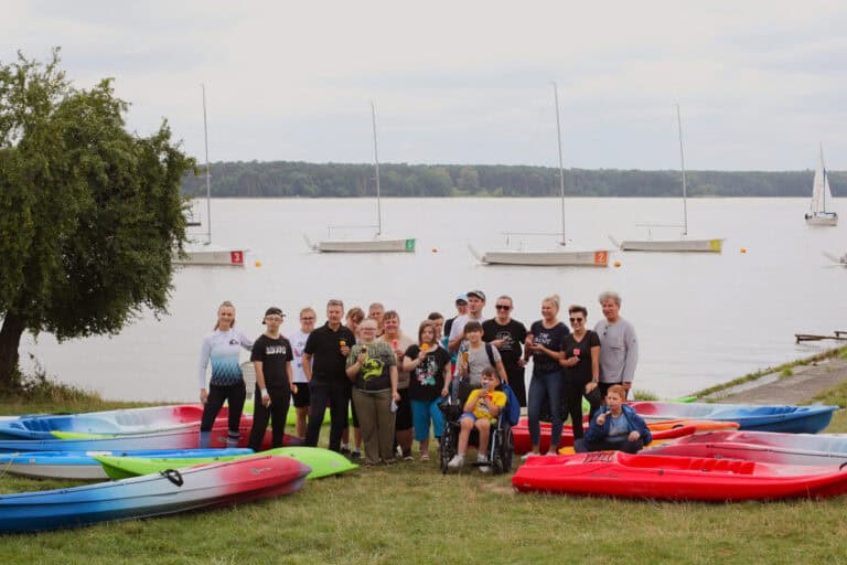 Na pierwszym planie między rozłożonymi na trawie kolorowymi kajakami stoi grupa kilkunastu osób pozujących do zdjęcia. Niektórzy trzymają w dłoniach lody na patyku. Przed nimi chłopiec na wózku. W tle zbiornik wodny i kilka żaglówek. Na horyzoncie linia lasu