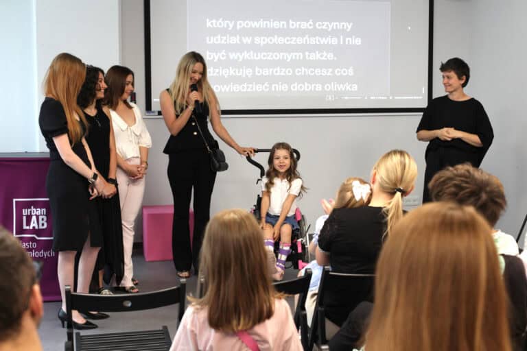 Zdjęcie z perspektywy tyłu sali, poprzez rząd ludzi siedzących na krzesłach. Drugi plan od lewej: cztery kobiety, z których jedna trzyma mikrofon, uśmiecha się. Druga dłoń spoczywa na rączce wózka, na którym siedzi mała uśmiechnięta dziewczynka. Z prawej tłumaczka języka migowego. W tle monitor z wyświetlanymi na żywo napisami