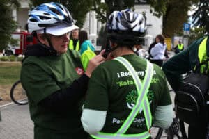 Na pierwszym planie dwie kobiety w kaskach i zielonych koszulkach. Kobieta z lewej pomaga zapiąć kask kobiecie stojącej tyłem do obiektywu. W tle zabudowania i zieleń drzew