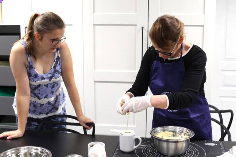 Dwie osoby: od lewej prowadząca zajęcia, przygląda się czynnościom, które wykonuje stojąca po prawej kobieta. Wbija ona jajko do białego kubka, używając specjalnego separatora do jaj. Za kobietami białe tło kuchennych szafek