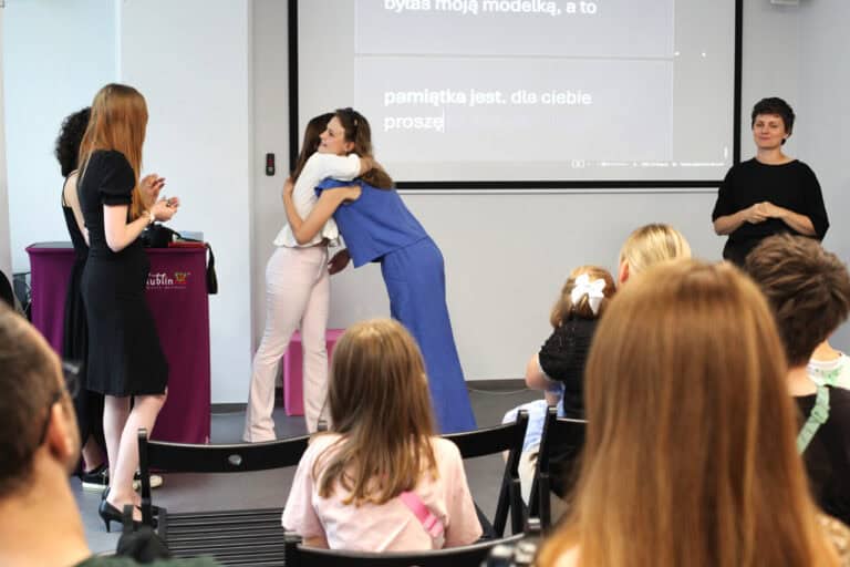 W centrum kadru dwie kobiety w uścisku. Po lewej: dwie klaszczące kobiety, które obserwują tą scenę. Po prawej tłumaczka języka migowego. W tle monitor z wyświetlanymi na żywo napisami. Zdjęcie wykonane z perspektywy tyłu sali, poprzez rząd ludzi siedzących na krzesłach