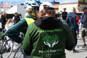 W centralnym punkcie kadru kobieta tyłem do obiektywu. Ma na sobie zieloną koszulkę z napisem: „Koła Dają Moc”, Pomoc Daje Moc. Fundacja”. W rozmytym tle kilka osób, budynki