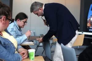 W kadrze od prawej siedząca kobieta. Prawą ręką wskazuje na ekran laptopa, który trzyma w ręce pochylony nad nią mężczyzna.