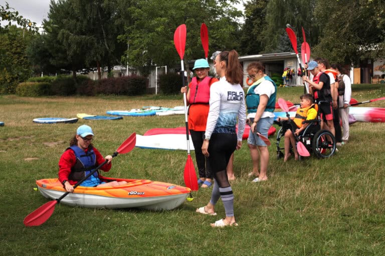 Na pierwszym planie młody mężczyzna w okularach i błękitnej czapce typu „kaszkietówka”, siedzi w kajaku i trzyma wiosło. Przed nim stoi instruktorka, która ma na sobie koszulkę z napisem na plecach "Fala Lublin". Obok nich kolejka stojących z wiosłami młodych ludzi