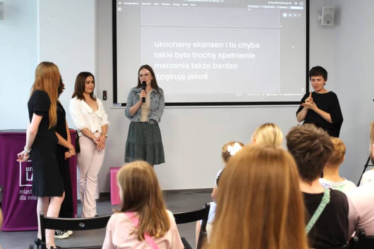 W centrum kadru od lewej cztery kobiety. Jedna z nich mówi do mikrofonu. Od prawej tłumaczka języka migowego. W tle monitor z wyświetlonym tekstem