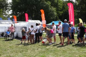 Od lewej kobieta na wózku, przy którym stoi mężczyzna. Obok nich namiot, przed którym tyłem do obiektywu stoi kolejka kilkunastu osób. W tle wysokie, podłużne, kolorowe chorągwie Jedynki Polskiego radia i zieleń drzew