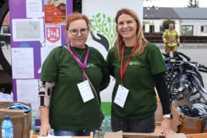 Pozowana fotografia. Dwie kobiety w zielonych koszulkach, z identyfikatorami na kolorowych smyczach. Za nimi banery z plakatem pikniku, trasą przejazdu i herbem Kocka