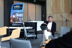 Od prawej banery stojące z napisem Lublin Gospodarczy i zdjęciem przeszklonego budynku. Z lewej siedzący na krześle mężczyzna, trzyma przed sobą, na kolanach otwarty laptop i wykonuje nad nim gest ręką. Za nim ściana z drewnianych paneli