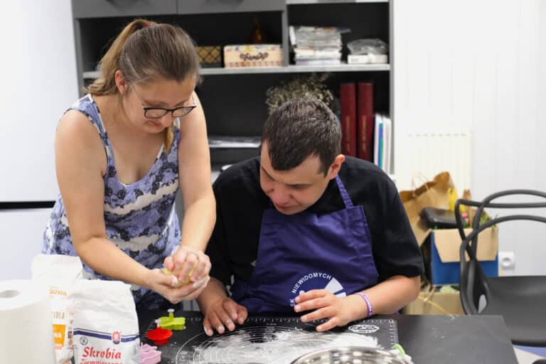 Dwie osoby: od lewej instruktorka, która w dłoniach ugniata ciasto. Przygląda się temu mężczyzna siedzący po prawej. Mężczyzna ma na sobie czarną koszulkę i granatowy fartuch. Na stole przed nim czarna mata do wyrabiania ciasta, oprószona mąką. W tle szary regał