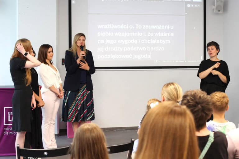 Pięć kobiet. Od lewej: dwie organizatorki, autorka fotografii Paulina Muszyńska, przy mikrofonie Zastępca Prezydenta ds. Kultury, Sportu i Partycypacji- Beata Stepaniuk-Kuśmierzak. W oddaleniu po prawej tłumaczka języka migowego. W tle ekran z wyświetlanym na żywo tekstem