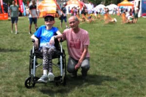 Centrum kadru - do fotografii pozują: młoda kobieta siedząca na wózku. Ubrana jest w getry, niebieską koszulkę z krótkim rękawem, czapkę z daszkiem, pozuje w ciemnych okularach. Kobieta uśmiecha się. Obok niej przykucnął mężczyzna, który jedną ręką trzyma podłokietnik wózka. W tle zieleń i rozmyty widok na namioty i ludzi