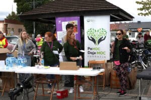 Cztery kobiety na tle banerów Fundacji Pomoc Daje Moc. Kobieta z prawej ma ciemne okulary i trzyma w ręku mikrofon. W tle zgromadzeni ludzie i dachy zabudowań