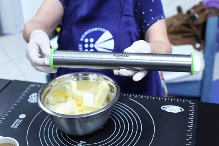 Zbliżenie na specjalny wałek do ciasta, z podziałką. Widać dłonie w jednorazowych rękawiczkach, które trzymają wałek nad metalową miską, w której znajduje się masło. Miska stoi na czarnej macie do wyrabiania ciasta