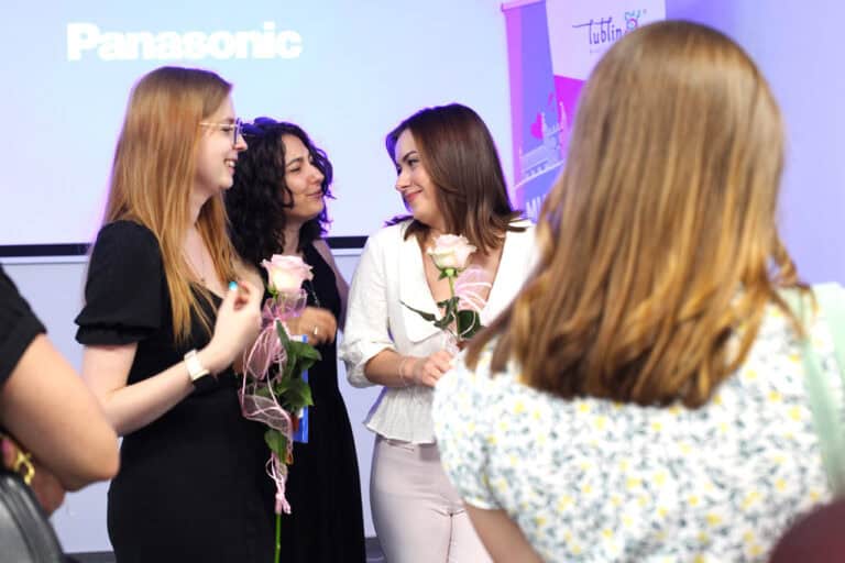 Trzy kobiety. Dwie z nich trzymają jasnoróżowe róże przewiązane różową wstążką. Kobiety uśmiechają się do siebie