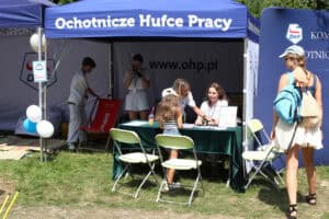 Granatowy namiot z napisem nad wejściem: Ochotnicze Hufce Pracy. W jego wnętrzu przy stole cztery osoby, przed stołem dziewczynka - tyłem do obiektywu. Po prawej stronie kadru młoda kobieta z plecakami, tyłem do obiektywu