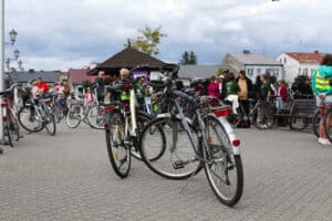 Na pierwszym planie dwa stojące na stopkach rowery. W tle zgromadzeni ludzie rowery, zabudowania. Niebo niebieskie, pochmurne