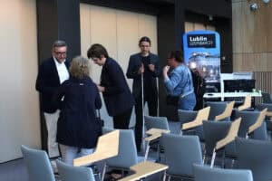 Na pierwszym planie, po przekątnej rzędy pustych krzeseł. Od lewej pięć osób stojących w dwóch grupach: dwie kobiety i mężczyzna oraz mężczyzna z białą laską i stojąca przy nim kobieta