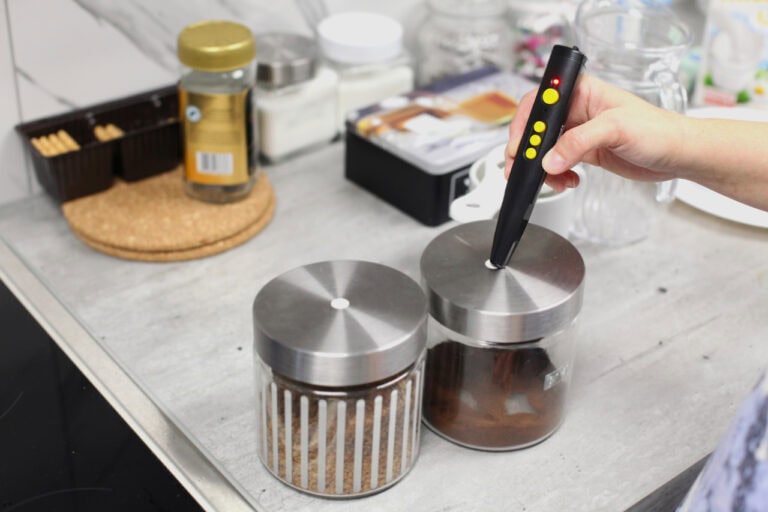 Na pierwszym planie szklane pojemniki z metalowymi nakrętkami i małymi, okrągłymi naklejkami, w których zaprogramowano nazwy produktów znajdujących się w środku. Widać dłoń z PenFriend – urządzeniem do głosowego etykietowania. W tle akcesoria kuchenne