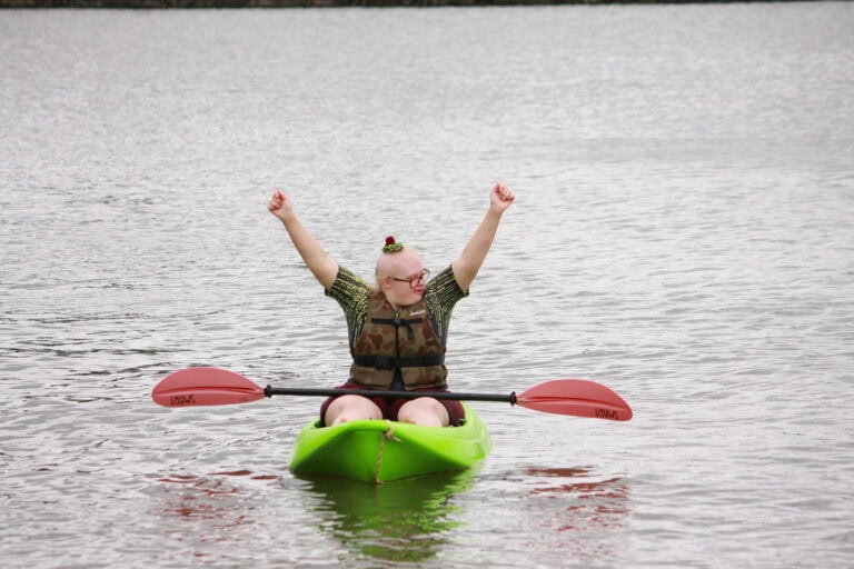 W centralnej części kadru na zielonym kajaku płynie młoda kobieta w okularach. Patrzy w bok unosząc w górę ręce. Na jej kolanach leży wiosło