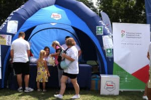 Od lewej granatowy namiot OHP, w środku i przed nim ludzie. Tuż obok namiotu duży stojący baner Państwowego Funduszu Rehabilitacji Osób Niepełnosprawnych