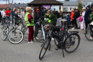 Na pierwszym planie czarny rower. W dalszej części kadru grupki ludzi zgromadzone przy rowerach. Niektórzy pogrążeni w rozmowie. W tle zabudowania
