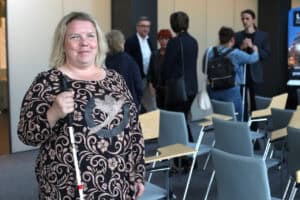 Do fotografii pozuje kobieta o jasnych włosach. Ma na sobie wzorzystą bluzkę w odcieniach brązu, w prawej ręce trzyma białą laskę. Kobieta uśmiecha się. Od lewej, w tle grupa osób pogrążonych w rozmowie