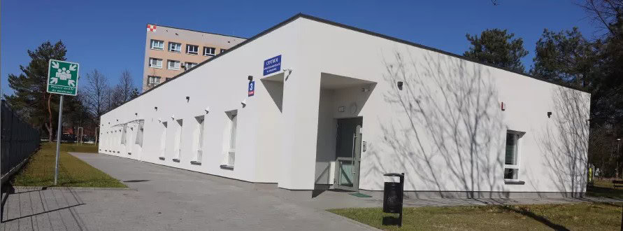Budynek biały parterowy, na tle większego, wielopiętrowego budynku mieszkalnego. Na drugim planie widoczny zielony znak drogowy oraz cienie rzucane przez pobliskie drzewa.