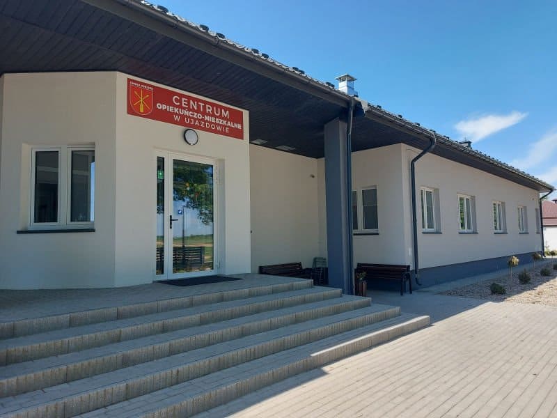 Parterowy budynek z tabliczką 'Centrum Opiekuńczo-Mieszkalne w Ujazdowie' nad wejściem. Do wejścia prowadzą schody, na zewnątrz ustawiono ławki.