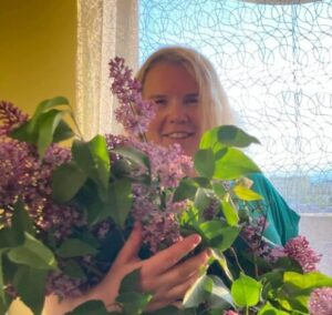 Uśmiechnięta Ewelina stoi na tle balkonowego okna. W rękach trzyma ogromny bukiet fioletowego bzu.
