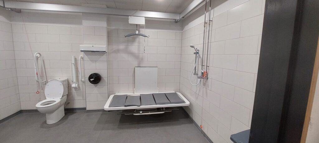 Wnętrze "Changing Places" na Lotnisku w Luton. Toaleta przystosowana dla osób niepełnosprawnych z sedesem, poręczami, przewijakiem montowanym na ścianie, podnośnikiem sufitowym, linkami awaryjnymi i prysznicem