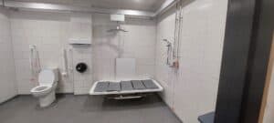 Wnętrze "Changing Places" na Lotnisku w Luton. Toaleta przystosowana dla osób niepełnosprawnych z sedesem, poręczami, przewijakiem montowanym na ścianie, podnośnikiem sufitowym, linkami awaryjnymi i prysznicem