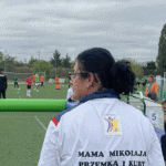 Kobieta w białej koszulce z napisem na plecach „mama Mikołaja, Przemka i Kuby” i Logo Fundacji Mały Krok Wielka Siła obserwuje zmagania piłkarzy na boisku.