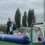 Fragment boiska w pobliżu bramki. Od lewej stoi trzyosobowa grupa rozmawiających, poniżej siedzi dwóch uśmiechniętych chłopców. W bramce stoi chłopiec.