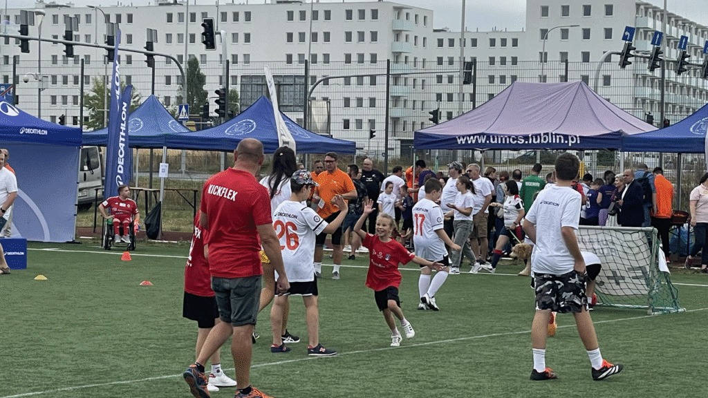 Grupa osób na boisku, od lewej mężczyzna w czerwonej koszulce z napisem KICKFLEX, chłopiec i uśmiechnięta dziewczynka przybijają „zwycięską” piątkę, inni zawodnicy na boisku, w tle pod parasolami UMCS, uczestnicy i kibice.