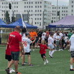 Grupa osób na boisku, od lewej mężczyzna w czerwonej koszulce z napisem KICKFLEX, chłopiec i uśmiechnięta dziewczynka przybijają „zwycięską” piątkę, inni zawodnicy na boisku, w tle pod parasolami UMCS, uczestnicy i kibice.