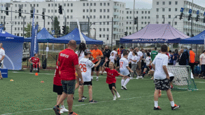 Grupa osób na boisku, od lewej mężczyzna w czerwonej koszulce z napisem KICKFLEX, chłopiec i uśmiechnięta dziewczynka przybijają „zwycięską” piątkę, inni zawodnicy na boisku, w tle pod parasolami UMCS, uczestnicy i kibice.