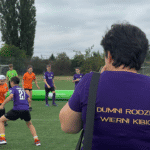 Od lewej strony, zawodnicy walczą o piłkę, w tle pozostali zawodnicy obserwują sytuację. Na pierwszym planie w prawej części fotografii, kobieta w fioletowej koszulce z napisem na plecach „DUMNI RODZICE WIERNI KIBICE”