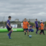 Zielona murawa boiska. Zawodnicy przy piłce, na boisku widoczni inni zawodnicy drużyn w pomarańczowych i fioletowych koszulkach