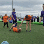 Boisko, zawodnicy w pomarańczowych i fioletowych koszulkach, dziewczyna podaje rękę leżącemu na boisku koledze z drużyny, chcąc pomóc mu wstać