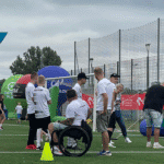 Zawodnicy i kibice w białych koszulkach stoją tyłem na murawie boiska. Jeden mężczyzna siedzi na wózku, u innego na koszulce widnieje nazwa „Proch Pionki”; w tle balony partnerów