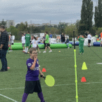 Chłopiec w fioletowym stroju sportowym, uśmiecha się, w ręku trzyma jaskrawożółtą neonową rakietkę, w tle uczestnicy wydarzenia