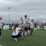 Grupa osób w koszulkach „Proch Pionki”, w centrum zainteresowania kobieta z dziewczynką pozujące do zdjęcia z łukiem, naprzeciwko nich klęczy kobieta ze smartfonem